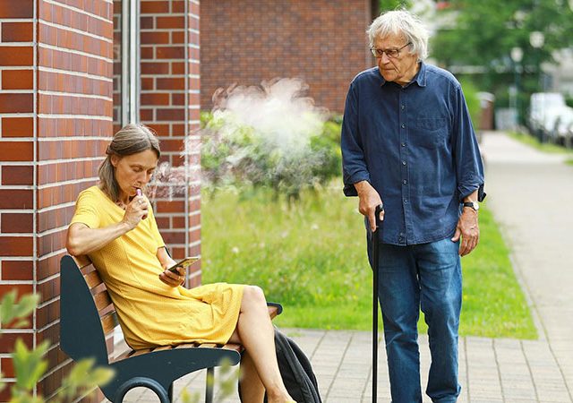 an elderly man standing near a woman smoking while sitting on a bench