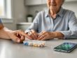 An older adult at a kitchen table with a smart pill organizer and a smartphone reminder, while a caregiver’s hand assists gently.